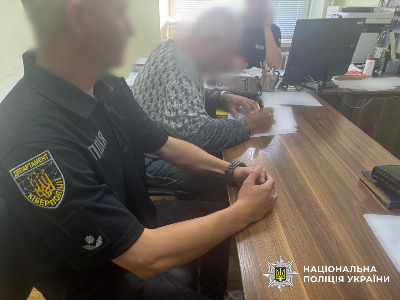 Працівник кіберполіції (в поліцейській формі з шевроном ДЕПАРТАМЕНТ КІБЕРПОЛІЦІЇ) сидить за дерев'яним столом біля обвинуваченого (чоловік у светрі). Обвинувачений підписує документ. Обличчя людей розмиті.