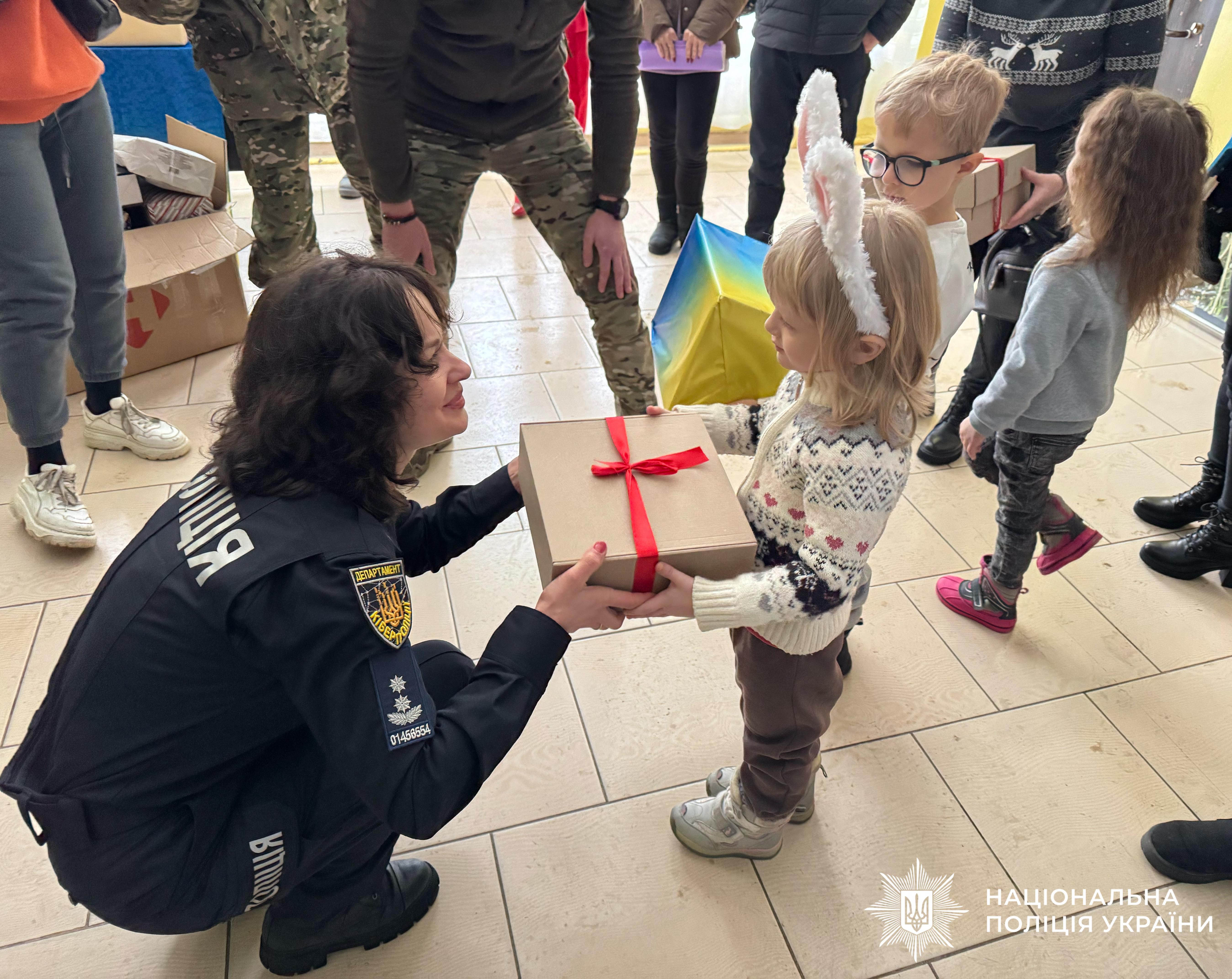 Жінка в чорній формі поліції з шевроном КІБЕРПОЛІЦІЯ присіла та передає картонну коробку з червоним бантом маленькій дівчинці в білому обідку з заячими вушками. Поруч стоїть хлопчик в окулярах, який тримає свій подарунок. На задньому плані видно інших дітей та військових у камуфляжній формі. У правому нижньому куті розміщено логотип Національної поліції України.