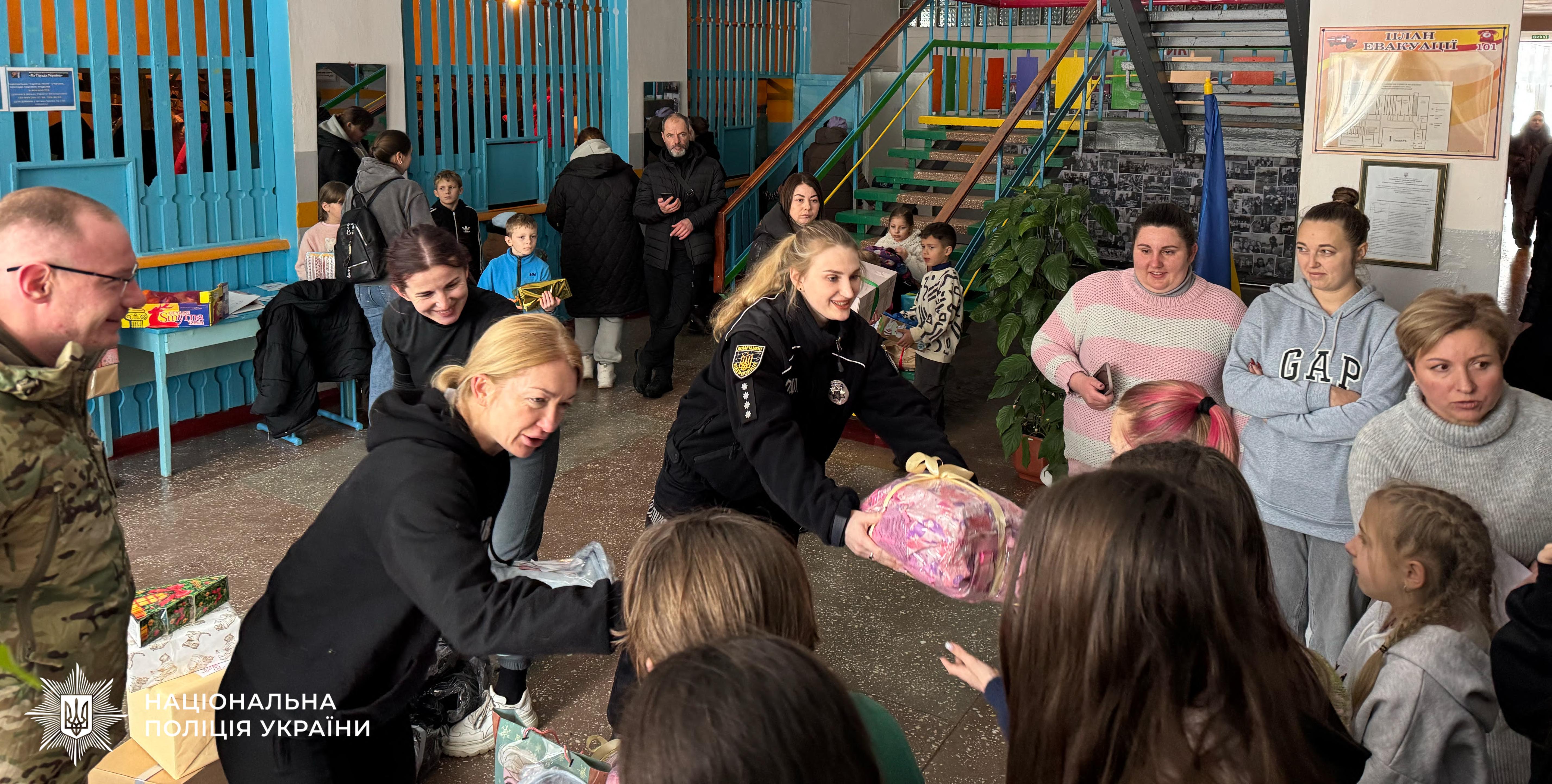 Фотографія у приміщенні, де група дорослих передає подарунки дітям. У центрі жінка у чорній формі поліції з шевроном Департаменту кіберполіції вручає дитині великий пакунок у рожевій обгортці. Поруч інша жінка у чорному худі також передає пакунок. Зліва стоїть усміхнений чоловік у військовій формі. На фоні видно інших людей, сходи та синю дерев'яну перегородку. У нижньому лівому куті розміщено логотип та напис: Національна поліція України.