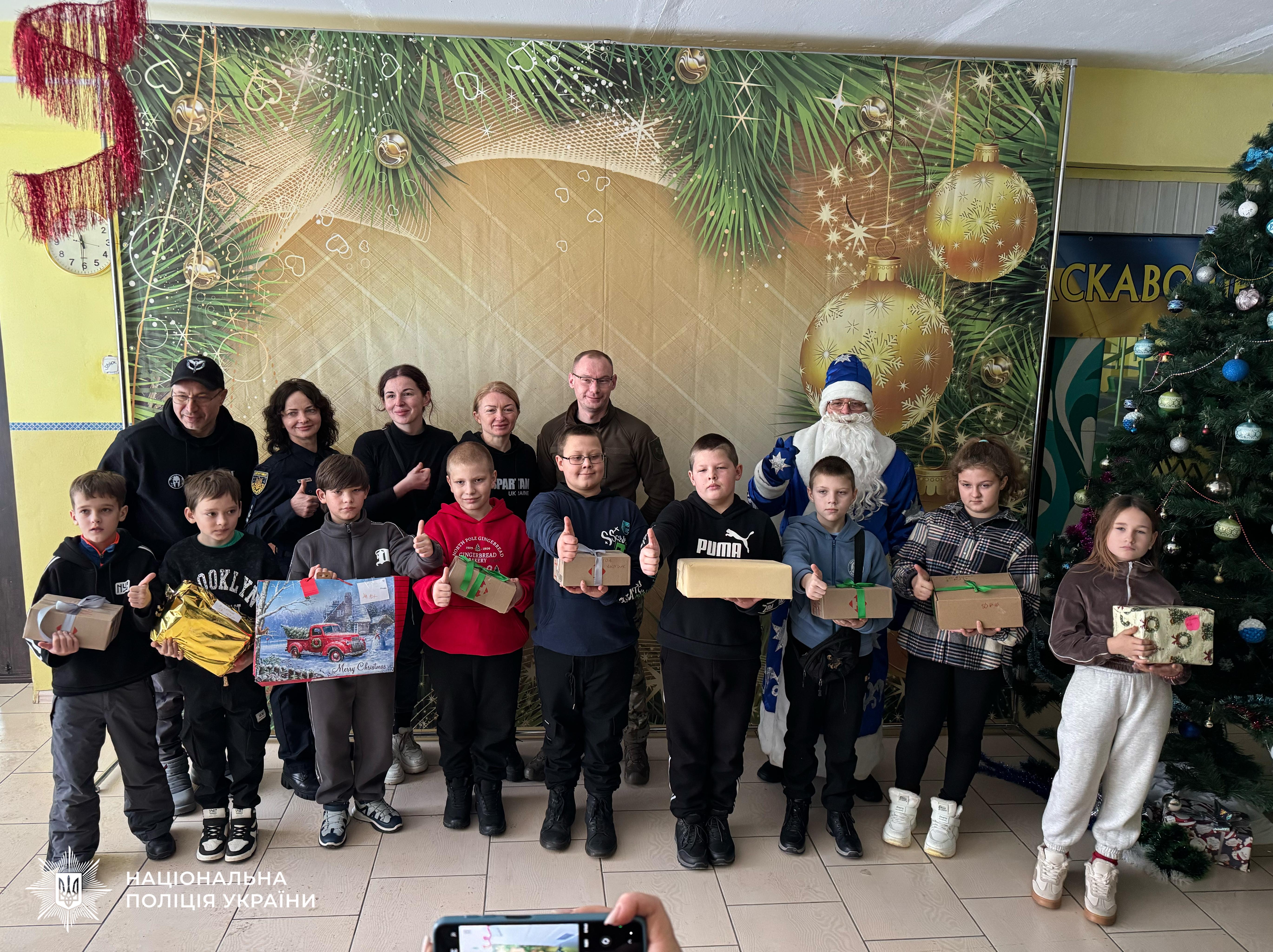 Групове фото дітей та дорослих у святковій залі. У передньому ряду стоять діти, які тримають у руках подарунки та показують жест «клас». За ними стоять дорослі, серед яких жінка у формі поліції, чоловік у військовій формі та аніматор у костюмі Святого Миколая у синьому вбранні. Праворуч стоїть прикрашена ялинка. На фоні розміщено великий святковий банер із зображенням ялинкових гілок та золотистих куль. У нижньому лівому куті розміщено логотип із текстом: Національна поліція України.