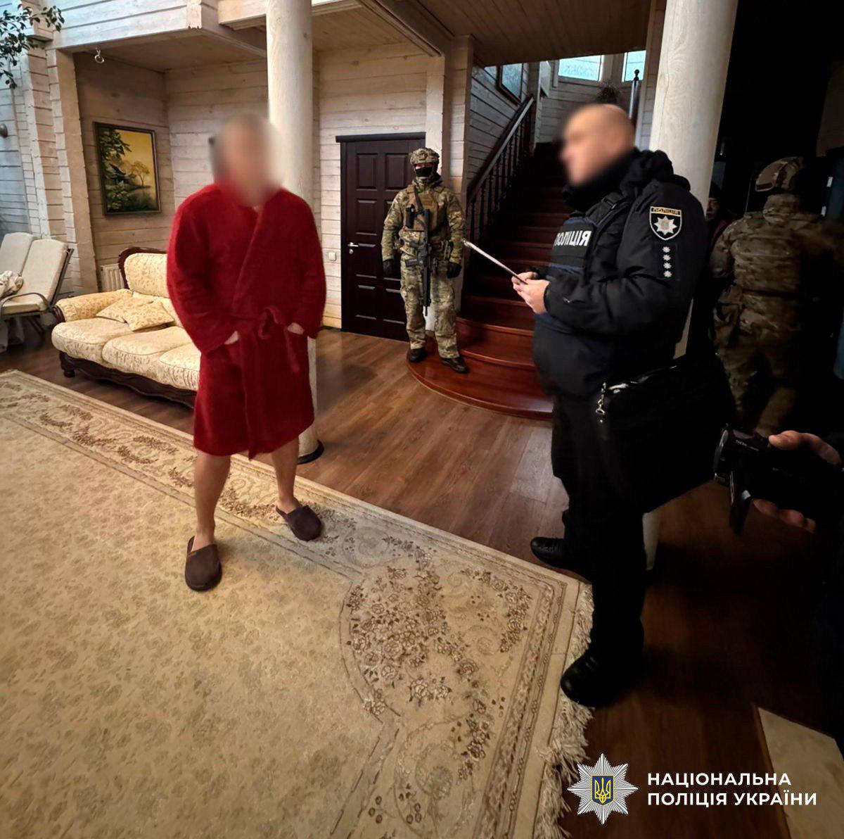 На фотографії зображено проведення слідчих дій у великій вітальні приватного будинку. Зліва стоїть чоловік у червоному махровому халаті та капцях, його обличчя розмито. Навпроти нього стоїть правоохоронець у чорній формі з написом Поліція на бронежилеті, який тримає в руках документи. На задньому плані біля сходів та дверей стоять двоє силовиків у повному камуфляжному спорядженні, шоломах та зі зброєю. Інтер'єр кімнати включає світлий диван, великий килим із візерунками та дерев'яні сходи на другий поверх. У нижньому правому куті розміщено логотип та напис Національна поліція України. Світлина ілюструє момент оголошення підозри або проведення обшуку.