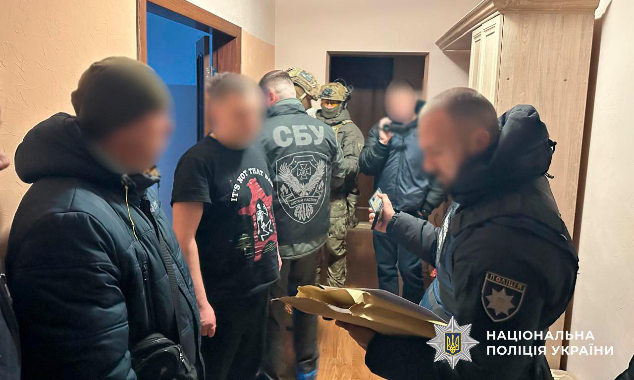 На фотографії зафіксовано проведення слідчих дій у передпокої житлового приміщення групою правоохоронців. На передньому плані справа стоїть поліцейський у чорній формі, який тримає розгорнуті документи та фіксує процес на мобільний телефон. У центрі кадру стоїть представник СБУ у тактичній куртці з великим написом СБУ на спині. Також присутні інші силовики у камуфляжі та балаклавах. Зліва стоять двоє чоловіків у цивільному одязі, їхні обличчя розмиті. У нижньому правому куті розміщено логотип та напис Національна поліція України. Світлина ілюструє спільну операцію різних підрозділів правоохоронних органів.