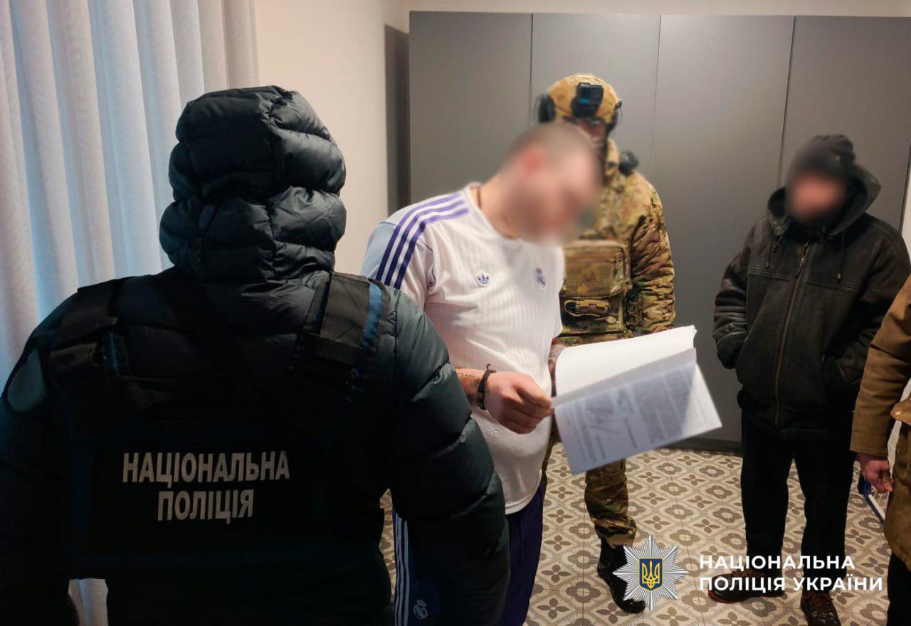 На фотографії зафіксовано момент вручення процесуальних документів під час слідчих дій у квартирі. На передньому плані зліва стоїть правоохоронець спиною до камери, на його куртці є напис Національна поліція. У центрі кадру чоловік у білій футболці уважно читає розгорнуті папери, його обличчя розмито. Поруч із ним стоїть силовик у камуфляжній формі, шоломі та захисних окулярах. На задньому плані видно ще одного чоловіка у темній куртці з капюшоном. Слідчі дії відбуваються у світлій кімнаті з сірою шафою та плиткою на підлозі. У нижньому правому куті розміщено логотип та напис Національна поліція України. Світлина ілюструє роботу поліції з ознайомлення підозрюваних із документами.