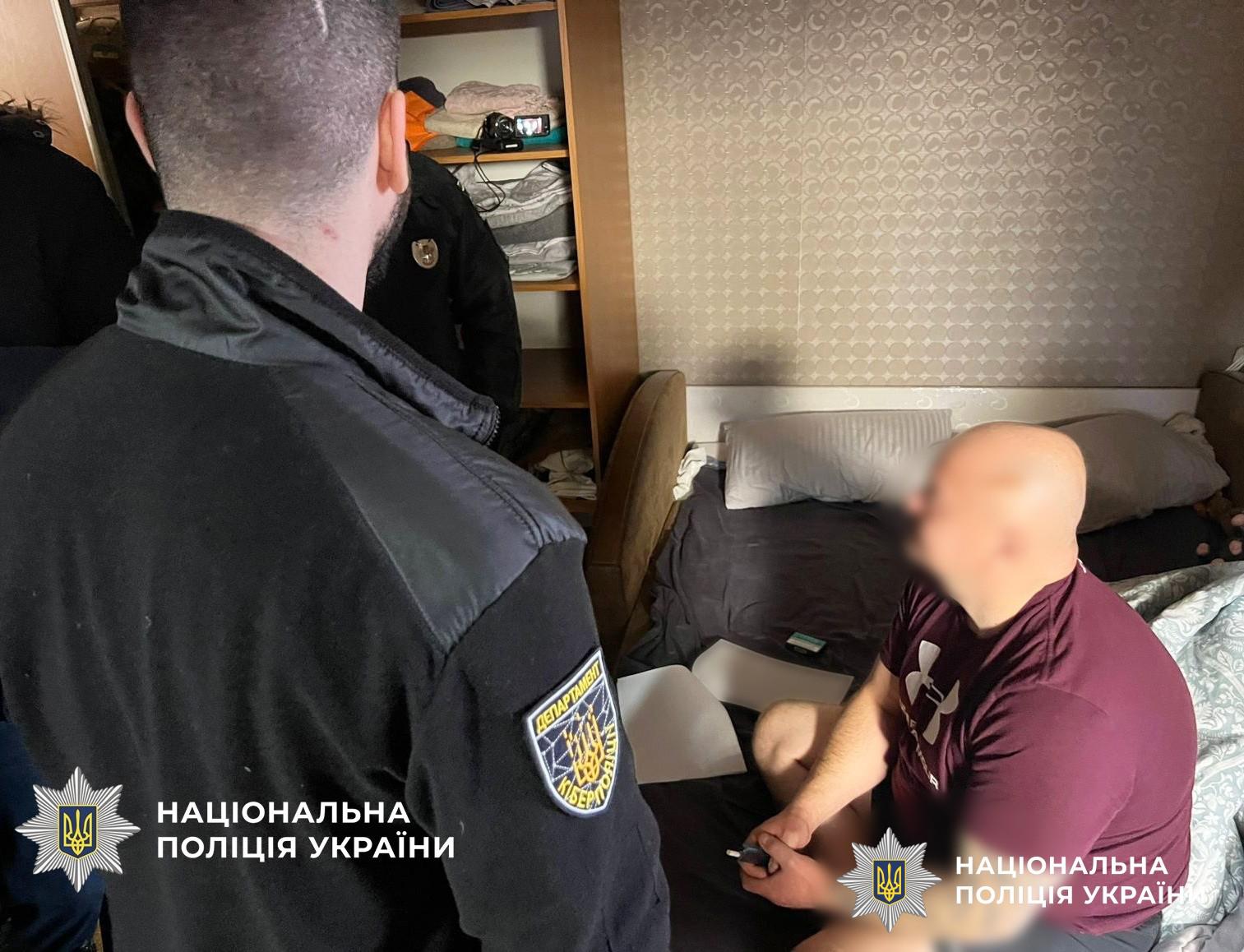 На фотографії зафіксовано проведення обшуку в житловій кімнаті. На передньому плані зліва стоїть правоохоронець у чорній формі з шевроном Департаменту кіберполіції, він повернутий спиною до камери. Навпроти нього на ліжку сидить чоловік у бордовій футболці, обличчя якого розмито. У кімнаті також видно частину дерев’яної шафи з речами та світлі шпалери на стінах. У нижніх кутах зображення розміщено логотипи та написи Національна поліція України. Світлина ілюструє роботу поліцейських під час слідчих дій у помешканні підозрюваного.