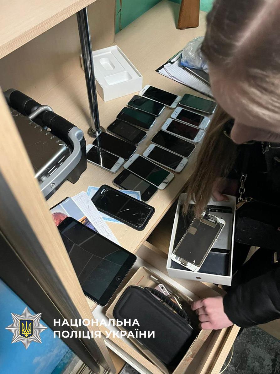  На фотографії зображено процес огляду речових доказів під час слідчих дій. На світлому дерев’яному столі та у відкритій шухляді розкладено велику кількість мобільних телефонів різних моделей. Більшість смартфонів мають чорні екрани, деякі з них пошкоджені або розібрані.  Поруч із телефонами лежить планшет, паперові документи та порожні коробки від техніки. Справа на знімку видно дівчину з довгим волоссям, яка перевіряє вміст однієї з коробок.  У лівому нижньому куті фотографії розміщено білий логотип Національної поліції України з відповідним написом. Зображення ілюструє вилучення технічних засобів, які могли використовуватися для незаконної діяльності.