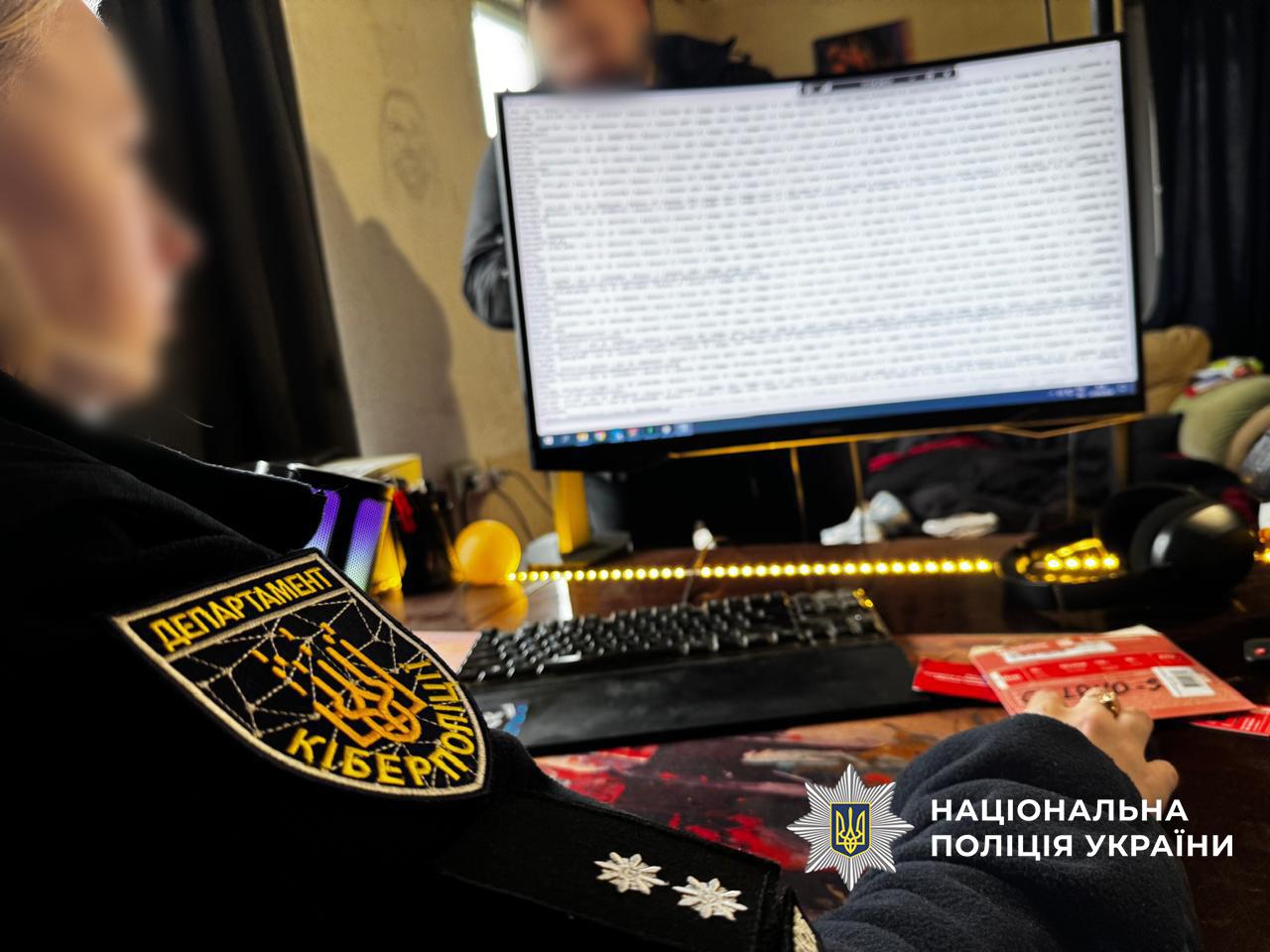 На фотографії зображено робоче місце під час проведення слідчих дій. На передньому плані зліва видно частину форми співробітника Департаменту кіберполіції. На рукаві — шеврон із гербом України та написом Департамент кіберполіції. На плечі помітні офіцерські погони з двома зірками.  У центрі кадру стоїть комп'ютерний монітор, на якому відкрито вікно з великим обсягом текстових даних, що нагадують програмний код або бази даних. Перед монітором лежить клавіатура. Стіл підсвічується жовтою світлодіодною стрічкою. На задньому плані видно розмиту постать людини, обличчя якої приховане.  У правому нижньому куті додано логотип Національної поліції України з відповідним написом білого кольору. Світлина зроблена в приміщенні з приглушеним освітленням.