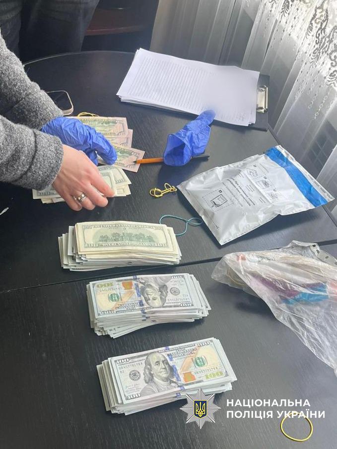 На фотографії зображено процес підрахунку та вилучення грошей під час слідчих дій. На темному столі розкладено кілька пачок готівки — купюри номіналом 100 доларів США. Зліва видно руки людини у блакитних гумових рукавичках, яка перераховує банкноти.  Також на столі лежать чисті аркуші паперу на планшеті, ручка, канцелярські гумки, прозорий поліетиленовий пакет та спеціальний білий пакет для речових доказів із маркуванням.  У нижньому правому куті фотографії розміщено білий логотип Національної поліції України з відповідним написом. Світлина ілюструє документування фінансових коштів, ймовірно здобутих незаконним шляхом.