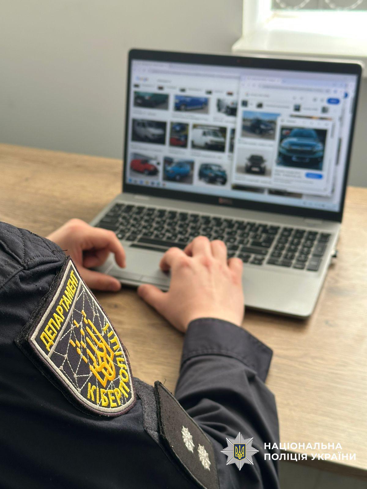На фотографії зображено співробітника поліції з боку спини та плеча, який працює за ноутбуком на дерев'яному столі. На рукаві чорної форми правоохоронця чітко видно шеврон Департаменту кіберполіції з жовтим тризубом на фоні стилізованої павутини та написом Департамент Кіберполіції. Руки людини лежать на клавіатурі ноутбука сірого кольору. На екрані відкрито браузер із результатами пошуку зображень різних автомобілів. У нижній правій частині кадру розміщено білий логотип Національної поліції України з написом Національна поліція України. Світло падає з вікна праворуч, створюючи природне освітлення робочого місця.