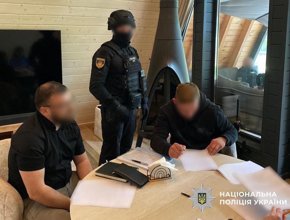 На фотографії зображено трьох осіб у світлій кімнаті з дерев'яним інтер'єром. За круглим світлим столом сидять двоє чоловіків у темному одязі, їхні обличчя розмиті. Чоловік праворуч схилився над білим аркушем паперу та пише ручкою, а чоловік ліворуч у чорній сорочці та окулярах сидить поруч і спостерігає. На столі також лежать закриті блокноти, папки з документами, ручка та серветниця. За ними стоїть правоохоронець у повному екіпіруванні: чорній формі, бронежилеті, шоломі та балаклаві, яка приховує обличчя. На його правому рукаві видно жовту нашивку Департаменту кіберполіції. На задньому плані розташовані великий телевізор, конусоподібний камін та панорамне вікно, у відображенні якого видно ще одного силовика. У правому нижньому куті кадру розміщено білий логотип Національної поліції України з відповідним підписом. Освітлення у приміщенні денне та природне.