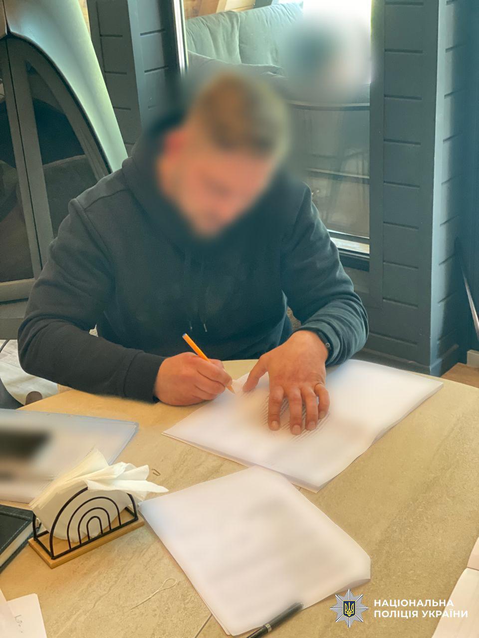 На фотографії зображено чоловіка у темно-синьому худі, який сидить за світлим столом у приміщенні та заповнює документи. Чоловік схилився над паперами, тримаючи в правій руці помаранчеву ручку, а лівою притримує аркуш; його обличчя розмито. На столі перед ним лежать кілька стопок паперів із друкованим текстом, деталі якого приховані розмиттям, а також стоїть металева серветниця з білими серветками та лежить чорна ручка. На задньому плані видно частину інтер'єру з темними вертикальними панелями та конусоподібною конструкцією каміна. У правому нижньому куті зображення розміщено білий логотип Національної поліції України з відповідним текстовим підписом. Знімок зроблений з ракурсу зверху під кутом, освітлення у кімнаті денне та рівномірне.
