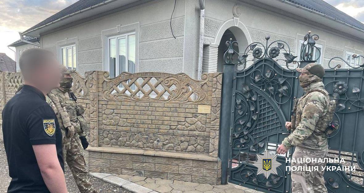На фотографії біля входу до приватного будинку зображено трьох людей. Зліва стоїть чоловік у цивільному, вдягнений у чорну футболку, на якій ззаду розташована жовто-чорна нашивка з гербом України та текстом «ДЕПАРТАМЕНТ» та «КІБЕРПОЛІЦІЯ». Поруч із ним перебуває чоловік у військовій камуфляжній формі та зі спорядженням, обличчя якого приховане балаклавою, повернутий спиною. Праворуч, біля металевих кованих воріт зеленого кольору із візерунком, стоїть ще один чоловік у камуфляжній формі та зі спорядженням, обличчя якого також закрите балаклавою. Обидва чоловіки в камуфляжі дивляться у бік воріт. На воротах у правому нижньому куті зображено логотип Національної поліції України та напис білими літерами: «НАЦІОНАЛЬНА ПОЛІЦІЯ УКРАЇНИ». Позаду людей знаходиться будинок зі світло-сірим фасадом та пластиковими вікнами, обгороджений комбінованим парканом із бетонних блоків коричневого та сірого кольорів із ажурною верхньою частиною. Небо на задньому плані світле.