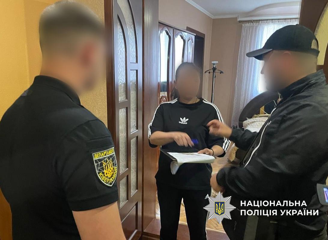 Зображення незаконий перетин кордону_1 копія.jpg показує інтер'єр приміщення, схожого на передпокій, де знаходяться три людини. На передньому плані ліворуч стоїть чоловік у чорній футболці-поло з нашивкою на рукаві, що містить герб та текст "ДЕПАРТАМЕНТ" і частково видимий напис "КІБЕР...". Обличчя чоловіка розмите. У центрі стоїть жінка, її обличчя також розмите. Вона одягнена в чорну футболку з білим логотипом "Adidas" на грудях та смугами на рукавах. Жінка дивиться вниз на папку-планшет із документами, які вона тримає в руках, і тримає ручку, ніби збирається підписати або робить позначки. Праворуч від неї стоїть третій чоловік у чорній куртці, чорній бейсболці та чорній масці-балаклаві, що закриває нижню частину обличчя. Його очі видно, але обличчя також розмите. Він повернутий напівбоком до жінки, жестикулює правою рукою, вказуючи на документи, які вона тримає. Між чоловіком ліворуч та жінкою видно дерев'яні міжкімнатні двері зі скляними вставками. За жінкою видно меблі (ймовірно, шафа із дзеркальними дверцятами), вішалку для одягу та частину іншої кімнати зі світлими стінами, вікном із прозорою шторою та, можливо, диваном. У правому нижньому куті зображення розміщено водяний знак із логотипом Національної поліції України та текстом білими літерами: "НАЦІОНАЛЬНА ПОЛІЦІЯ УКРАЇНИ". Зображення кольорове, освітлене звичайним світлом приміщення.