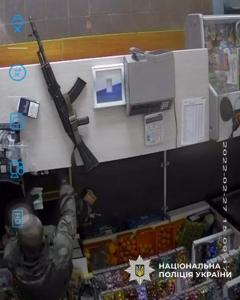 Зображення з камери спостереження всередині магазину, зняте з верхнього ракурсу. У нижній лівій частині видно людину у військовій камуфляжній формі, яка стоїть спиною до об'єктива біля прилавка та протягує руку до внутрішньої зони каси. На світлій поверхні прилавка лежить автомат Калашникова, поруч із яким розташовані касовий апарат, калькулятор, паперовий аркуш із таблицею, пачка цигарок та невелика фоторамка. У нижній частині кадру видно ящики з цитрусовими фруктами та полиці з іншими товарами. Уздовж правого краю кадру вертикально нанесено дату та час: 2022-02-27 15:09:47. На лівому краї зображення присутні напівпрозорі блакитні іконки інтерфейсу відеоплеєра. У нижньому правому кутку поверх кадру накладено офіційний білий логотип-зірку з гербом-тризубом на синьому тлі та білий напис Національна поліція України.