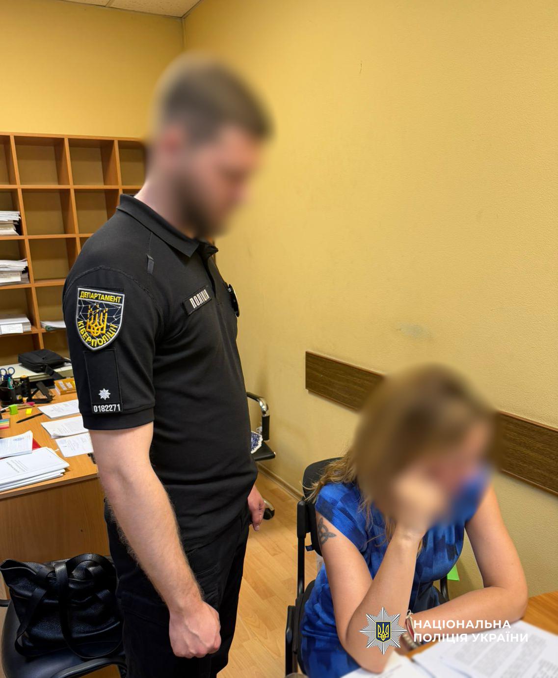 Працівник кіберполіції у формі стоїть у службовому кабінеті біля робочого столу. Поруч сидить жінка. Обличчя чоловіка та жінки приховані розмиттям. Поліцейський вдягнений у чорну футболку з нашивкою "Департамент кіберполіції" на рукаві. Жінка в блакитній футболці. У кабінеті видно офісні меблі, документи на столі та стелаж із паперами на задньому плані. У нижньому правому куті розміщено логотип "Національна поліція України".