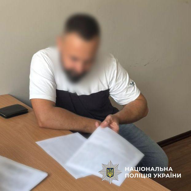 Фотографія у світлому приміщенні, на якій за дерев'яним столом сидить чоловік у двоколірній біло-чорній футболці та синіх джинсах. Його обличчя повністю розмите. Чоловік нахилився вперед, спершись правою рукою на стіл, і гортає пальцями лівої руки білі паперові документи, що лежать перед ним. Текст на паперах також розмитий. На лівому краю столу лежить невеликий чорний гаманець або блокнот, а поруч у лівому кутку видно куточок іншої розмитої папки з документами. Стіни кімнати однотонні світло-сірого кольору, підлога дерев'яна коричнева. У правому нижньому кутку поверх фотографії накладено білий логотип-зірку з гербом-тризубом на синьому тлі та білий напис Національна поліція України.