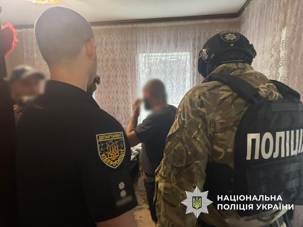 Світлина знята зсередини кімнати з візерунчастими шторами на вікні, де знаходяться кілька осіб. На передньому плані ліворуч спиною до об'єктива стоїть чоловік у чорній футболці із жовтим шевроном Департаменту кіберполіції та двома сріблястими зірками на рукаві. Праворуч від нього спиною до глядача стоїть правоохоронець у військовому камуфляжі, захисному шоломі та тактичному жилеті з великим білим написом ПОЛІЦІЯ на спині. У центрі на задньому плані стоїть чоловік у темній футболці, який тримає руку біля свого обличчя. Ліворуч на задньому плані видніється ще одна постать у кепці. Обличчя людей на задньому плані повністю розмиті. У правому нижньому кутку поверх фотографії накладено білий логотип-зірку з гербом-тризубом на синьому тлі та білий напис Національна поліція України.