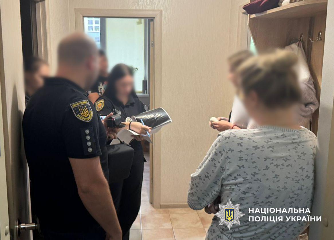 Світлина зсередини житлового приміщення, де в коридорі перебувають кілька людей. На передньому плані ліворуч напівбоком до глядача стоїть чоловік у чорній футболці із шевроном Департаменту кіберполіції на правому рукаві та чотирма зірками на погоні. Він тримає лівою рукою відеокамеру. Далі у глибині коридору видно жінку у чорній формі, яка робить записи ручкою на аркуші паперу. Навпроти них праворуч спиною до глядача стоїть жінка у світлій домашній кофті з візерунком у вигляді метеликів, яка тримає схрещені на грудях руки. Біля неї стоїть ще одна людина у білому одязі. Усі обличчя людей на фотографії повністю розмиті. Стіни приміщення світло-бежеві, підлога викладена плиткою, праворуч помітно дерев'яну вішалку для одягу. У правому нижньому кутку поверх фотографії накладено білий логотип-зірку з гербом-тризубом на синьому тлі та білий напис Національна поліція України.