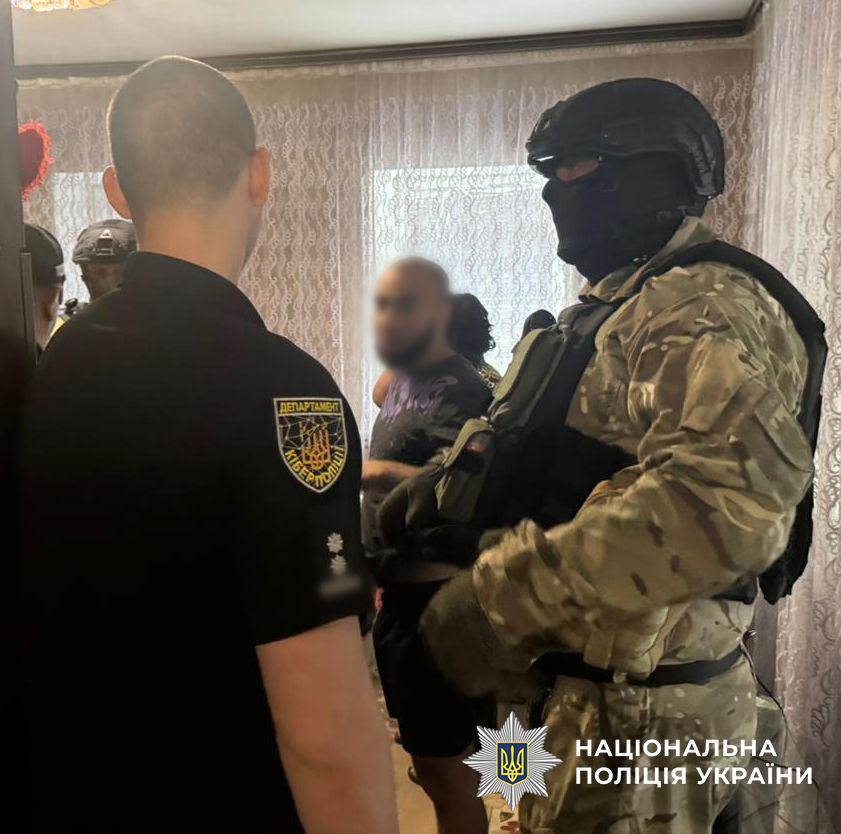 Світлина з інтер'єру кімнати зі світлими візерунчастими шторами на вікні. На передньому плані ліворуч спиною до об'єктива стоїть чоловік у чорній футболці, на правому рукаві якої помітно жовтий круглий шеврон Департаменту кіберполіції. Навпроти нього праворуч стоїть правоохоронець у військовому камуфляжі, захисному шоломі, тактичному жилеті та чорній балаклаві, що повністю закриває обличчя. Посередині на задньому плані стоїть бородатий чоловік у темній футболці з принтом, його обличчя розмите. У правому нижньому кутку поверх фотографії накладено білий логотип-зірку з гербом-тризубом на синьому тлі та білий напис Національна поліція України.