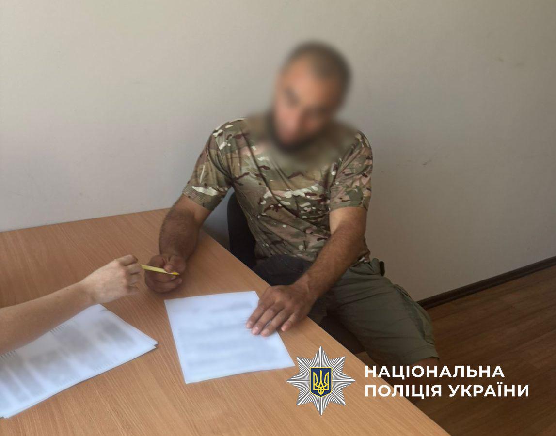 Фотографія у світлому приміщенні, де за дерев'яним столом на стільці сидить чоловік у камуфляжній футболці та зелених шортах. Його обличчя повністю розмите. Чоловік тримає жовту ручку в правій руці та спирається лівою рукою на білий аркуш паперу, що лежить перед ним на столі. Текст на цьому папері розмитий. З лівого боку в кадр потрапляє рука іншої людини, яка тримає ручку та розташована поруч із ще одним стосом розмитих документів. Стіни кімнати однотонні та світлі, підлога дерев'яна коричневого кольору. У правому нижньому кутку поверх фотографії накладено білий логотип-зірку з гербом-тризубом на синьому тлі та білий напис Національна поліція України.