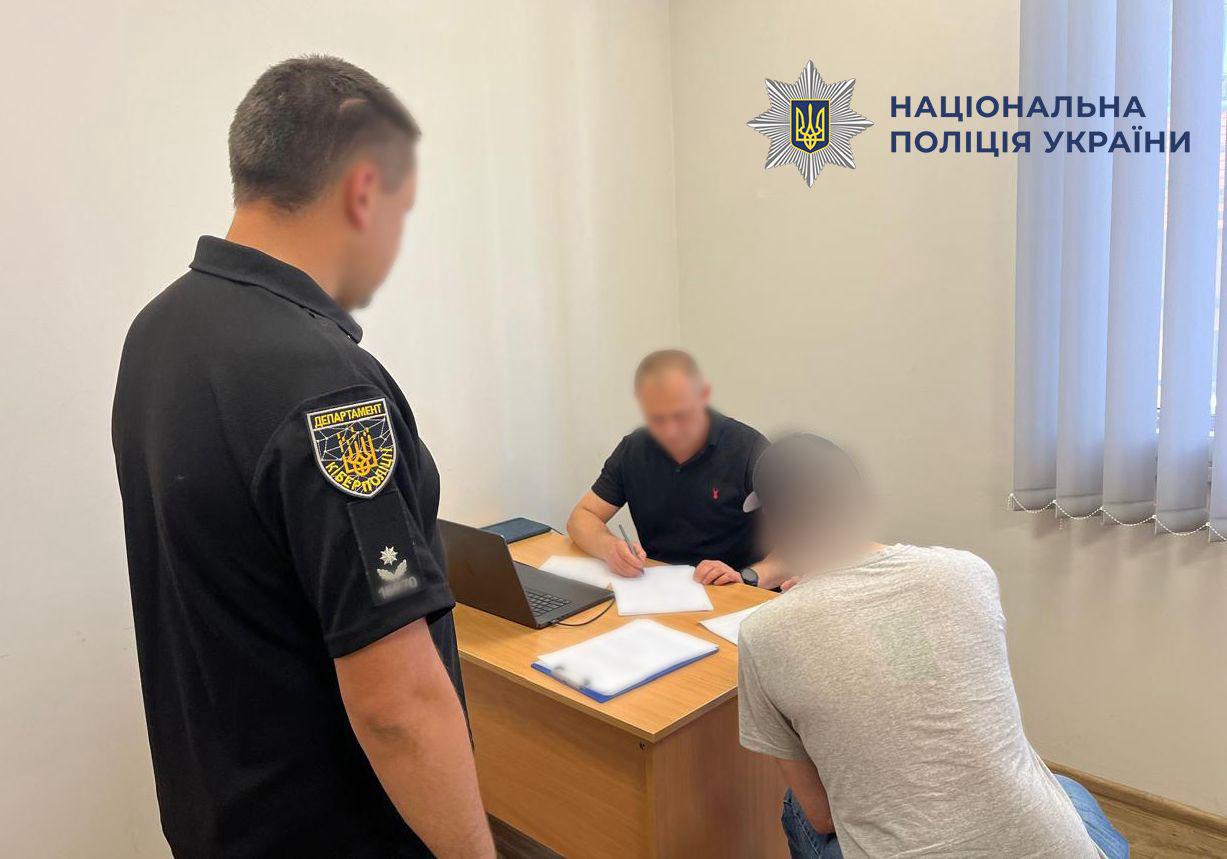 Фотографія у світлому офісному приміщенні, де троє людей перебувають біля робочого столу. На передньому плані ліворуч спиною до об'єктива стоїть чоловік у чорній футболці-поло з жовтим шевроном Департаменту кіберполіції та сріблястою зіркою на лівому рукаві. На передньому плані праворуч спиною до глядача сидить людина у світло-сірій футболці та синіх джинсах. У центрі за дерев'яним столом сидит чоловік у чорній футболці, який тримає ручку та робить записи на білих аркушах паперу. На столі поруч із паперами та синьою папкою стоїть відкритий темний ноутбук. Обличчя усіх трьох людей повністю розмиті. Стіни кімнати білі, праворуч на вікні висять світлі вертикальні жалюзі. У верхньому правому кутку поверх фотографії накладено білий логотип-зірку з гербом-тризубом та темний напис Національна поліція України.