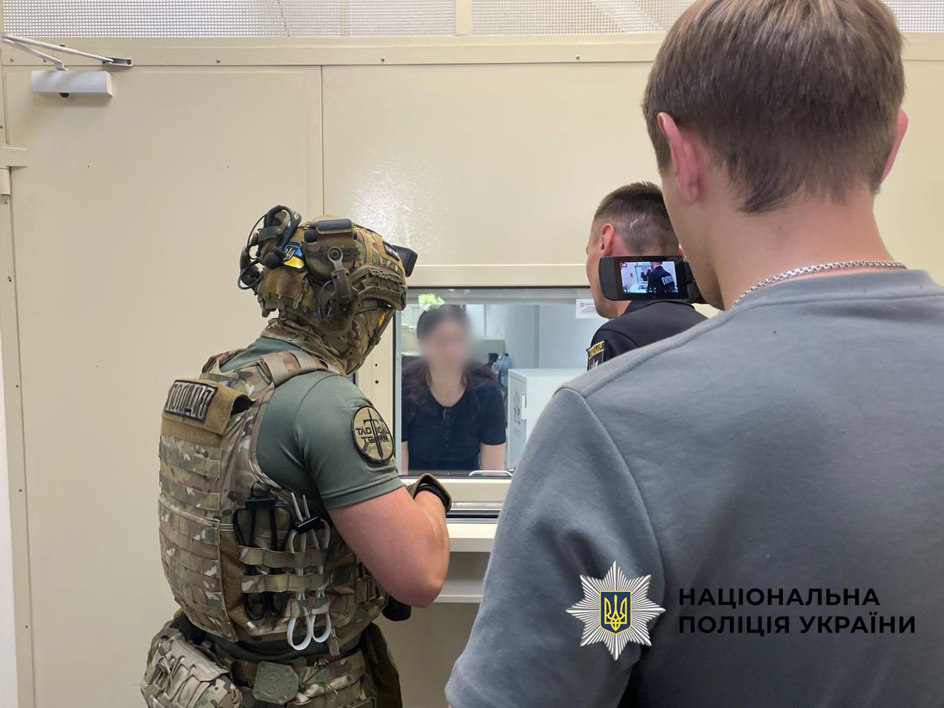Світлина у приміщенні зі світло-бежевими стінами, де кілька людей стоять перед прозорим віконцем. Ліворуч спиною до глядача стоїть правоохоронець у військовому камуфляжному жилеті, зеленій футболці, шоломі з маленьким прапором України та балаклаві. На його лівому рукаві є кругла нашивка з написом TACTICAL TEAM. У центрі через скляне віконце видно темноволосу жінку в чорній футболці, обличчя якої повністю розмите. Поруч із віконцем стоїть чоловік у темній формі, який знімає її на невелику відеокамеру. На передньому плані праворуч спиною до об'єктива стоїть чоловік у сірій футболці та зі срібним ланцюжком на шиї. У правому нижньому кутку поверх фотографії накладено білий логотип-зірку з гербом-тризубом на синьому тлі та чорний напис Національна поліція України.
