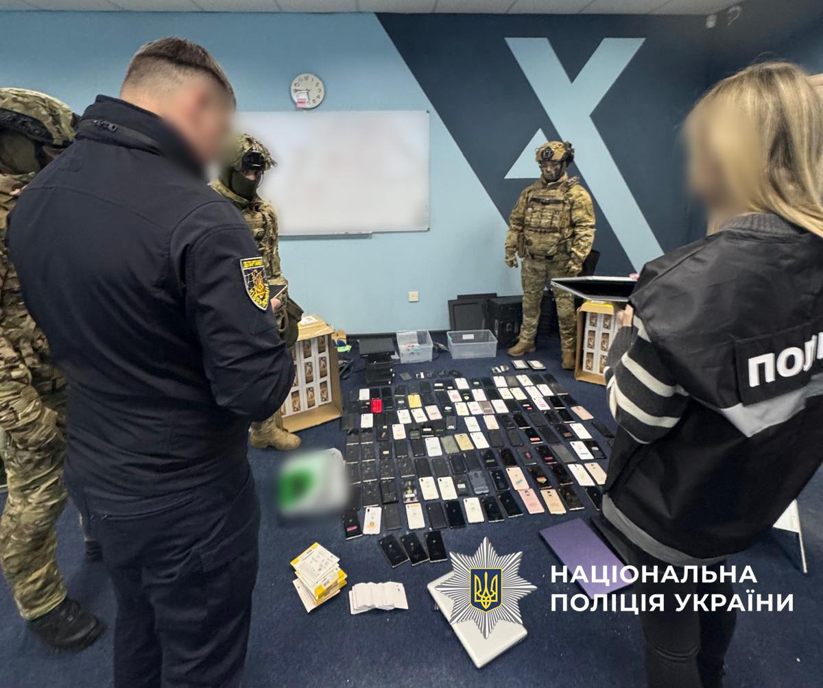 Світлина у приміщенні з блакитними та темно-синіми стінами, де на підлозі розкладено численні мобільні телефони, планшети, ноутбуки, пачки SIM-карт та картонні коробки. Навколо техніки стоять кілька людей: чоловік у темно-синьому куртці з шевроном Департаменту кіберполіції на рукаві, жінка праворуч у жилетці з білим написом ПОЛІЦІЯ, яка тримає в руках планшет, а також троє осіб у військовому камуфляжі, захисних шоломах та балаклавах. Обличчя цивільних людей на фотографії розмиті. На задній стіні висить біла дошка та видно фрагмент великого світлого логотипа у формі літери X. У нижній частині по центру накладено білий логотип-зірку з гербом-тризубом на синьому тлі та білий напис Національна поліція України.