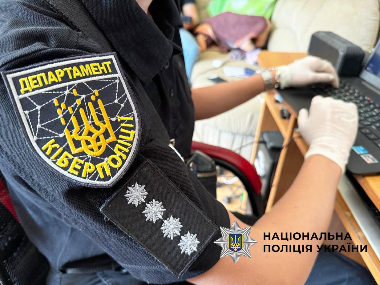 Фотографія знята з близької відстані та показує людину в чорній формі. На її лівому рукаві чітко видно шеврон Департаменту кіберполіції. Шеврон має форму щита чорного кольору з жовтою окантовкою, на якому жовтими літерами написано ДЕПАРТАМЕНТ КІБЕРПОЛІЦІЇ та зображено жовтий герб-тризуб, стилізований під елементи цифрової мережі. Нижче на рукаві прикріплений погон із чотирма сріблястими восьмикутними зірками, розміщеними в ряд. Людина сидить у кріслі та тримає руки в білих гумових рукавичках на клавіатурі відкритого темного ноутбука, що стоїть на дерев'яному столі. На задньому плані видно розмиті обриси житлової кімнати з м'якими меблями. У правому нижньому кутку поверх фотографії накладено білий логотип-зірку з гербом-тризубом на синьому тлі та білий напис НАЦІОНАЛЬНА ПОЛІЦІЯ УКРАЇНИ.