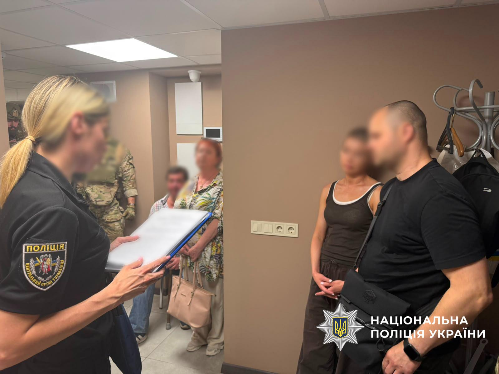 Світлина у приміщенні з бежевими стінами, де перебувають кілька осіб. На передньому плані ліворуч напівбоком до глядача стоїть жінка з білявим волоссям у чорній поліцейській формі з шевроном ПОЛІЦІЯ ЦЕНТРАЛЬНИЙ ОРГАН УПРАВЛІННЯ на рукаві. Вона тримає в руках папку-планшет із паперами, текст яких розмитий. На передньому плані праворуч стоять чоловік у чорній футболці із сумкою через плече та жінка у темній майці. На задньому плані видно ще кількох людей: сивого чоловіка, жінку у візерунчастій блузці з сумкою на лікті, а також двох осіб у військовому камуфляжі. Обличчя більшості присутніх цивільних осіб розмиті. Справа біля стіни стоїть підлогова вішалка з одягом, а на стіні видно розетки та вимикачі. У правому нижньому кутку поверх фотографії накладено білий логотип-зірку з гербом-тризубом на синьому тлі та білий напис Национальна поліція України.