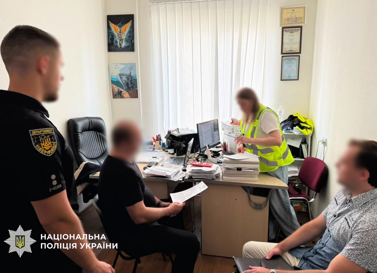 Світлина в робочому кабінеті, де перебувають чотири особи. На передньому плані ліворуч стоїть чоловік у чорній поліцейській формі з шевроном Департаменту кіберполіції на рукаві. У центрі за дерев'яним столом, заваленим паперами та папками, сидить чоловік у чорній футболці та тримає в руках аркуш паперу. За цим же столом стоїть жінка у білій футболці та яскраво-жовтому світловідбиваючому жилеті, яка перебирає документи біля монітора комп'ютера. На передньому плані праворуч сидить чоловік у короткорукавній сорочці. Обличчя всіх осіб розмиті. На білій стіні висять два картини-малюнки та три грамоти в рамкаx, а вікно завішене білими жалюзі. У лівому нижньому кутку поверх фотографії накладено білий логотип-зірку з гербом-тризубом на синьому тлі та білий напис Національна поліція України.