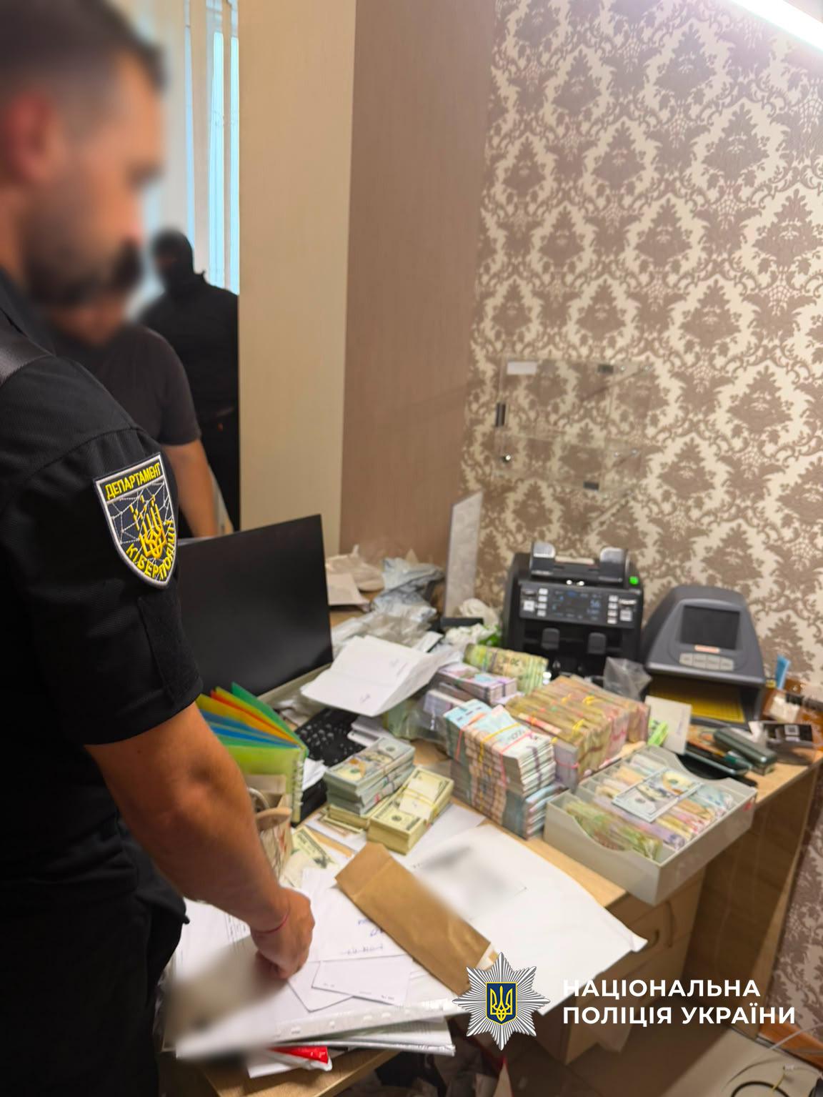 Світлина в приміщенні з візерунчастими шпалерами, де біля столу стоїть чоловік у чорній поліцейській формі з шевроном Департаменту кіберполіції на рукаві. Його обличчя розмите. На передньому плані видно дерев'яний стіл, завалений паперами, конвертами та високими стосами пачок грошових купюр у гривнях та доларах США, перетягнутих гумками. На столі також стоїть відкритий лоток з грошима, монітор, клавіатура та дві лічильні машинки. На задньому плані в проймі дверей видно ще двох осіб, одна з яких у чорній балаклаві. У правому нижньому кутку поверх фотографії накладено білий логотип-зірку з гербом-тризубом на синьому тлі та білий напис Національна поліція України.