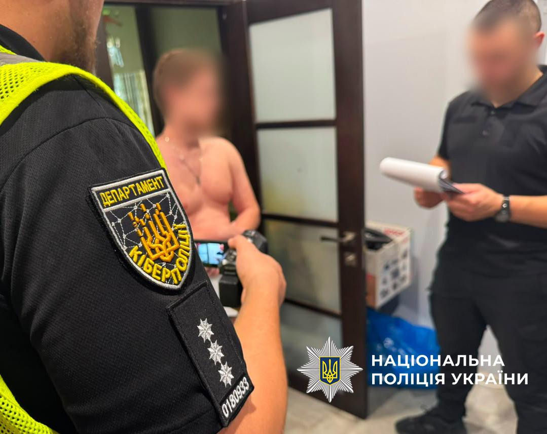 Світлина у приміщенні біля дверного отвору, де перебувають кілька людей. На передньому плані ліворуч спиною до об'єктива стоїть правоохоронець у чорній футболці зі світловідбиваючим жилетом та шевроном Департаменту кіберполіції на рукаві. Він тримає в руках невелику відеокамеру і здійснює зйомку. У центрі на задньому плані стоїть чоловік із оголеним торсом, обличчя якого розмите. Праворуч стоїть чоловік у чорній сорочці поло та чорних штанях, який тримає в руках білий блокнот із паперами і дивиться в нього; його обличчя також розмите. У правій нижній частині поверх фотографії накладено білий логотип-зірку з гербом-тризубом на синьому тлі та білий напис Національна поліція України.