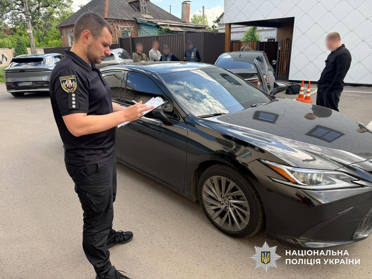 Чоловік у чорній поліцейській формі з шевроном ПОЛІЦІЯ стоїть зліва біля передньої частини чорного легкового автомобіля на асфальтованій парковці та пише ручкою в блокноті. Праворуч від автомобіля, спиною до об'єктива, стоїть інший чоловік у чорному одязі біля дорожніх конусів, його обличчя розмите. На задньому плані, біля паркану та будівель, стоять ще п'ятеро людей, їхні обличчя також розмиті. Видно приватні житлові будинки: один старий одноповерховий з темно-сірим шиферним дахом і один сучасний двоповерховий з білим фасадом, а також паркани і дерева. На парковці стоїть ще один сірий позашляховик. На асфальті біля автомобілів є тіні. У нижньому правому куті на зображенні розміщено логотип: біла зірка з гербом і синім текстом НАЦІОНАЛЬНА ПОЛІЦІЯ УКРАЇНИ.