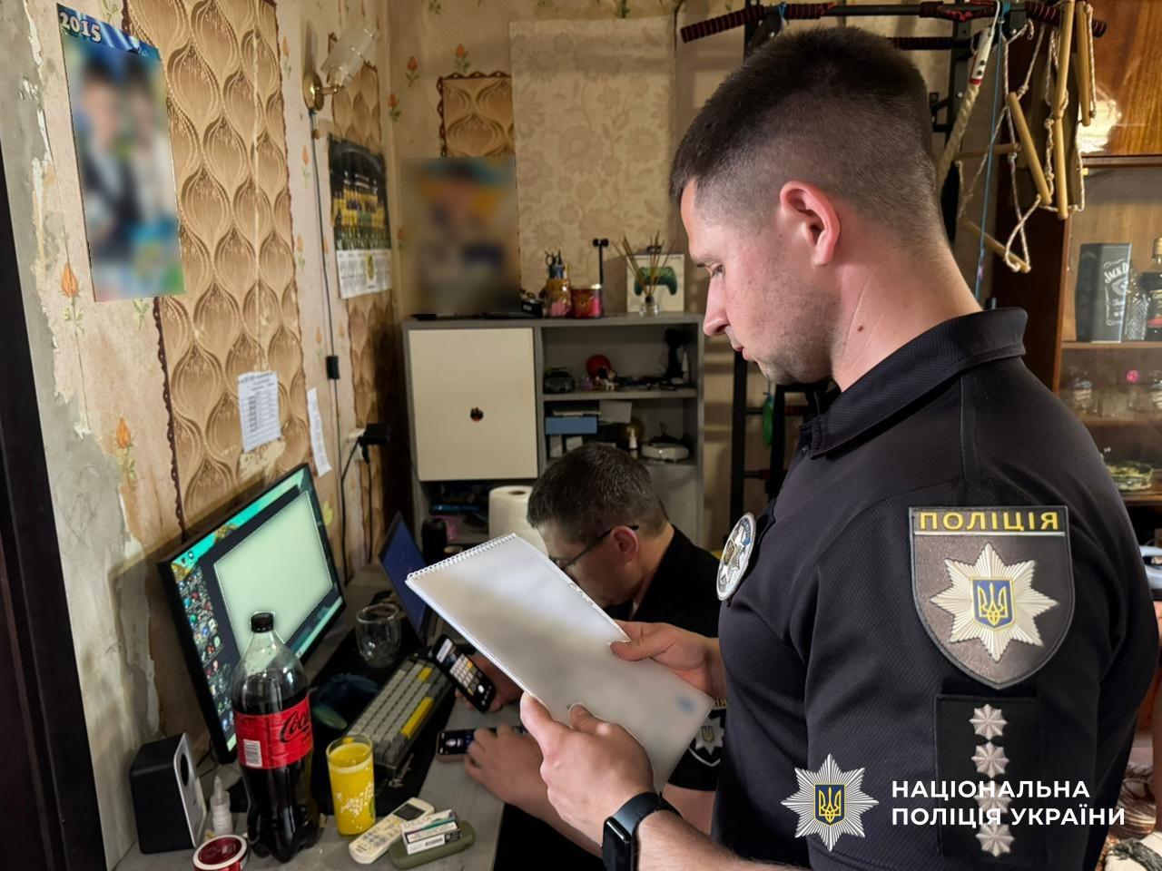 Світлина у житловій кімнаті зі старими візерунчастими шпалерами. На передньому плані праворуч у профіль стоїть правоохоронець у чорній поліцейській формі з шевроном ПОЛІЦІЯ на рукаві. Він тримає у руках білий блокнот зі спіраллю та дивиться в нього. Поруч із ним за комп'ютерним столом сидить інший чоловік в окулярах, який працює з мобільними телефонами та ноутбуком. На столі біля увімкненого монітора комп'ютера стоять пляшка напою Coca-Cola, склянка з жовтим напоєм, клавіатура та лежать дрібні речі. На стіні ліворуч висять плакати з розмитими зображеннями. У правому нижньому кутку поверх фотографії накладено білий логотип-зірку з гербом-тризубом на синьому тлі та білий напис Національна поліція України.