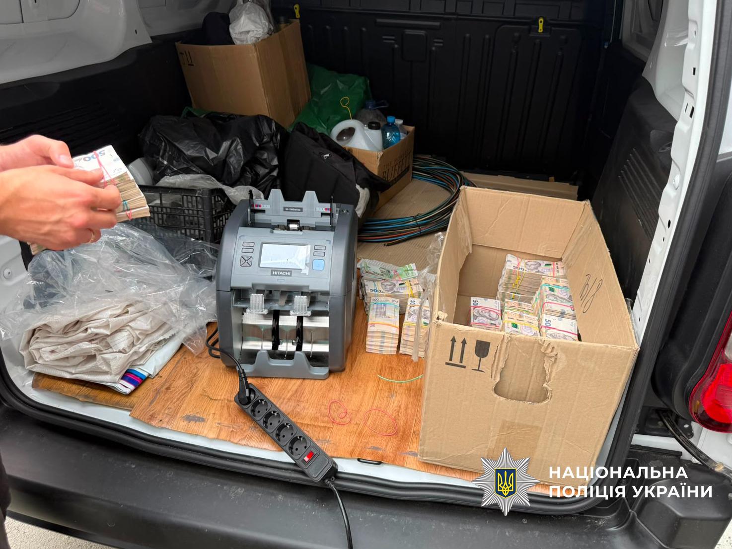Світлина відкритого багажника автомобіля, всередині якого розкладені речі. У центрі стоїть сіра електронна лічильна машинка для грошей, підключена до чорного подовжувача. Праворуч розташована велика картонна коробка, заповнена пачками купюр гривень різного номіналу, перетягнутими гумками. Ще кілька пачок грошей лежать поруч із коробкою та лічильною машинкою. Ліворуч чиїсь руки тримають стопку банкнот номіналом п'ятсот гривень. На задньому плані багажника видно картонні коробки, пакети, пластикові пляшки та згорнуті дроти. У правому нижньому кутку поверх фотографії накладено білий логотип-зірку з гербом-тризубом на синьому тлі та білий напис Національна поліція України.