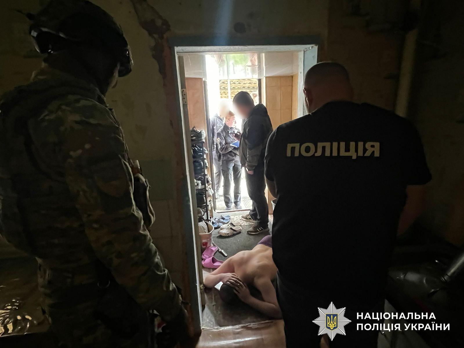 Світлина у напівтемному приміщенні біля відчинених дверей до передпокою. На передньому плані ліворуч спиною до об'єктива стоїть силовик у камуфляжній формі та шоломі. Праворуч спиною стоїть правоохоронець у чорній футболці з великим написом ПОЛІЦІЯ білого кольору на спині. На підлозі біля дверей обличчям донизу лежить чоловік із оголеним торсом, тримаючи руки біля голови. У коридорі за дверима, освітленому денним світлом, стоять троє осіб: одна людина в чорній куртці, обличчя якої розмите, та жінка в поліцейській формі з блокнотом у руках. У передпокої видно полицю з взуттям та капці на підлозі. У правому нижньому кутку поверх фотографії накладено білий логотип-зірку з гербом-тризубом на синьому тлі та білий напис Національна поліція України.