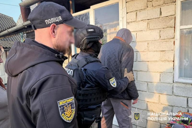 Світлина біля цегляної стіни будівлі на подвір'ї. На передньому плані ліворуч стоїть правоохоронець у чорній кепці з білим написом ПОЛІЦІЯ та в чорній куртці із шевроном ДЕПАРТАМЕНТ КІБЕРПОЛІЦІЇ на рукаві, його обличчя розмите. У центрі стоїть силовик у тактичному шоломі, балаклаві та бронежилеті, який тримає за плече чоловіка, повернутого обличчям до стіни. Чоловік біля стіни одягнений у темну стьобану куртку з накинутим капюшоном та сірі штани. Поруч видно скляні двері та вікно будинку. У правому нижньому кутку поверх фотографії накладено білий логотип-зірку з гербом-тризубом на синьому тлі та білий напис НАЦІОНАЛЬНА ПОЛІЦІЯ УКРАЇНИ.