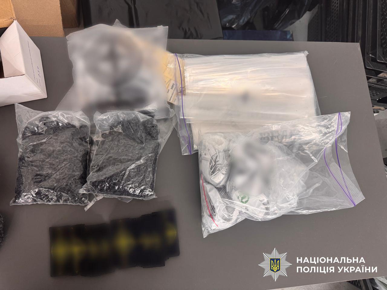 Світлина предметів, розкладених на сірій горизонтальній поверхні. На передньому плані ліворуч лежать два прозорі пластикові пакети з зип-застібкою, наповнені дрібними чорними ґудзиками, а нижче них розташовані прямокутні темні заготовки чи деталі, зображення яких розмите. Праворуч на поверхні лежить великий прозорий пакет із зип-застібкою, у якому знаходяться білі паперові етикетки та бирки для одягу, частина з яких також розмита. У верхній частині фотографії розміщено напівпрозорий матовий пакет та стопку прозорих упаковок. У лівому верхньому кутку видно край білої картонної коробки. У правому нижньому кутку поверх зображення накладено білий логотип-зірку з гербом-тризубом на синьому тлі та білий напис НАЦІОНАЛЬНА ПОЛІЦІЯ УКРАЇНИ.