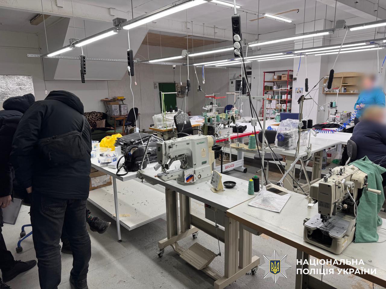 Світлина у світлому виробничому приміщенні швейного цеху. На передньому плані та вглиб кімнати розташовані декілька рядів білих робочих столів зі швейними машинами, котушками ниток різних кольорів, тканиною та інструментами. Понад столами на стелі закріплені довгі світлодіодні світильники та звисають електричні подовжувачі. У лівій частині фотографії спиною до камери стоять двоє чоловіків у темному верхньому одязі, які нахилилися над столом із чорними пакетами та особистими речами. У правій частині приміщення на задньому плані сидять дві людини, їхні обличчя розмиті, а біля стіни видно червоний стелаж із матеріалами. У правому нижньому кутку поверх фотографії накладено білий логотип-зірку з гербом-тризубом на синьому тлі та білий напис НАЦІОНАЛЬНА ПОЛІЦІЯ УКРАЇНИ.