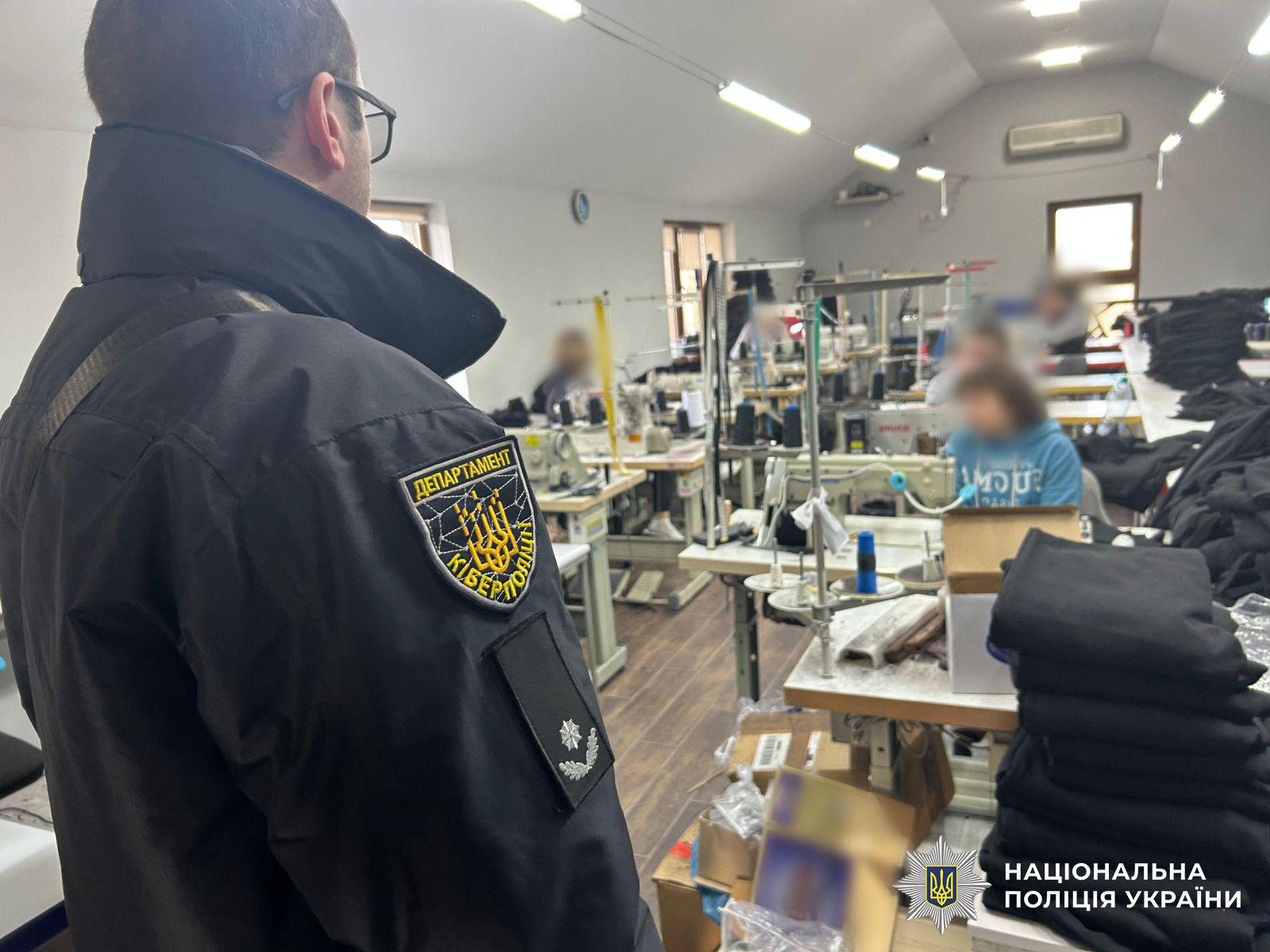 Світлина у світлому просторому приміщенні швейного цеху. На передньому плані зліва спиною та в профіль стоїть чоловік в окулярах та чорній куртці із шевроном ДЕПАРТАМЕНТ КІБЕРПОЛІЦІЇ на рукаві. На задньому плані вздовж кімнати розташовані робочі столи зі швейними машинами, котушками ниток, коробками та стопками згорнутої чорної тканини. За столами сидять кілька людей, їхні обличчя розмиті. У приміщенні горять довгі люмінесцентні лампи на стелі, а на стіні видно вікна та кондиціонер. У правому нижньому кутку поверх фотографії накладено білий логотип-зірку з гербом-тризубом на синьому тлі та білий напис НАЦІОНАЛЬНА ПОЛІЦІЯ УКРАЇНИ.