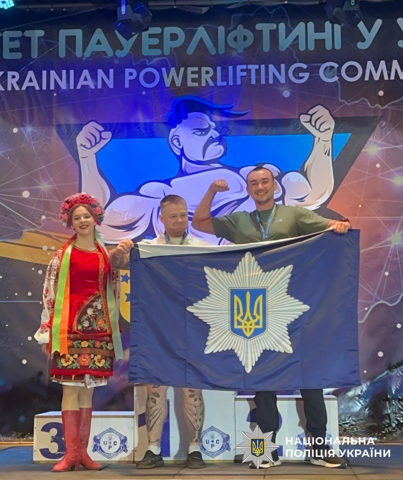 Світлина з змагань з пауерліфтингу, де люди розгортають прапор. У центрі кадру двоє чоловіків-спортсменів та дівчина у традиційному українському костюмі тримають великий темно-синій прапор із емблемою Національної поліції України — восьмикутною зіркою та гербом-тризубом у центрі. Чоловік у зеленій футболці та чорних штанах, який стоїть на п'єдесталі під номером один, посміхається і піднімає зігнуту в лікті руку, демонструючи біцепс. Ліворуч від нього стоять другий спортсмен у білій футболці та дівчина у червоній вишитій сукні з віночком і стрічками на голові. На тлі розміщено баннер із синьо-жовтою символікою та ілюстрацією козака-пауерліфтера з зігнутими руками, а також текстом УКРАЇНСЬКИЙ ПАУЕРЛІФТИНГ. У правому нижньому кутку поверх фотографії накладено білий логотип-зірку з гербом-тризубом на синьому тлі та білий напис НАЦІОНАЛЬНА ПОЛІЦІЯ УКРАЇНИ.