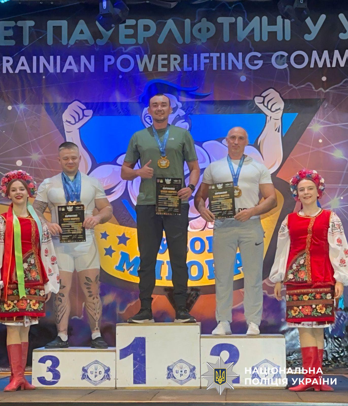 Світлина з церемонії нагородження на змаганнях з пауерліфтингу. На п'єдесталі пошани стоять троє чоловіків-спортсменів із медалями на шиях та сертифікатами в руках. На першому місці по центру стоїть чоловік у зеленій футболці та темних штанах, який посміхається і показує жест «великий палець угору». На другому місці праворуч стоїть лисий чоловік у білій футболці та сірих спортивних штанах. На третьому місці ліворуч стоїть чоловік у білій футболці з татуюваннями на ногах. По обидва боки від п'єдесталу стоять дві дівчини в українських національних костюмах із віночками на головах. На задньому плані розміщено синьо-жовтий банер із написом УКРАЇНСЬКИЙ ПАУЕРЛІФТИНГ та ілюстрацією м'язистої фігури. У правому нижньому кутку поверх фотографії накладено білий логотип-зірку з гербом-тризубом на синьому тлі та білий напис НАЦІОНАЛЬНА ПОЛІЦІЯ УКРАЇНИ.