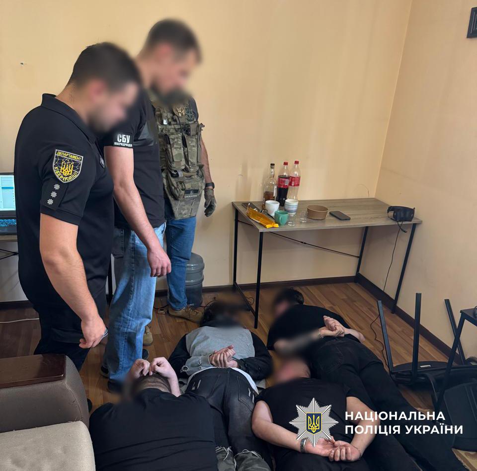 Фотографія у приміщенні, де на дерев'яній підлозі лежать обличчям донизу чотири чоловіки з заведеними за спину руками, обличчя яких розмиті. Біля них стоять троє правоохоронців: один у чорному поло з шевроном Департаменту кіберполіції, другий у чорній футболці з написом СБУ, а третій у тактичному жилеті камуфляжного забарвлення. Усі обличчя присутніх також розмиті. У кутку кімнати стоїть дерев'яний стіл, на якому розміщені пляшки, чашки та дрібні предмети. У правому нижньому кутку зображення міститься білий логотип та напис Національна поліція України.