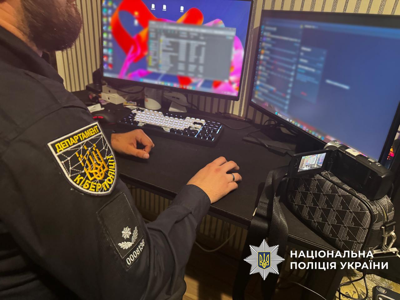 Фотографія, на якій чоловік у формі співробітника Департаменту кіберполіції з відповідним шевроном на рукаві сидить за робочим столом перед двома комп'ютерними моніторами. Його руки лежать на столі біля клавіатури та миші. На екранах моніторів видно відкриті інтерфейси програм та робочий стіл. На столі праворуч від комп'ютера стоїть невелика відеокамера на штативі. У правому нижньому кутку зображення розміщено білий логотип та напис Національна поліція України.