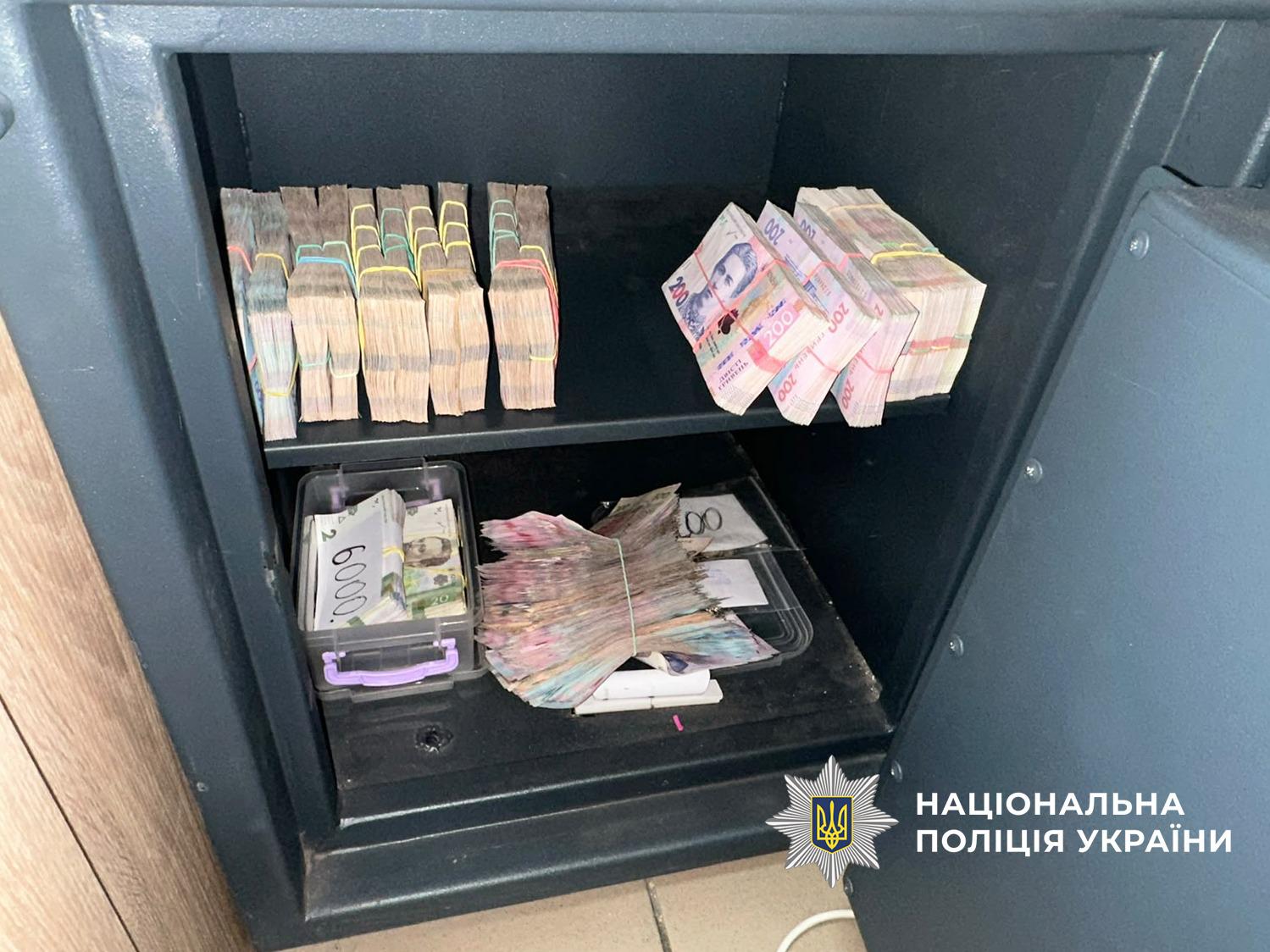 Фотографія відкритого металевого сейфа темно-сірого кольору, поділеного полицею на дві секції. На верхній полиці ліворуч стоять вертикально кілька товстих пачок паперових купюр, скріплених канцелярськими гумками, а праворуч лежать кілька пачок купюр номіналом двісті гривень. На нижній полиці розташовано прозорий пластиковий контейнер із купюрами, на якому написане число 6000, а поруч лежать ще дві пачки паперових грошей, скріплені гумками. У правому нижньому кутку зображення розміщено білий логотип та напис Національна поліція України.